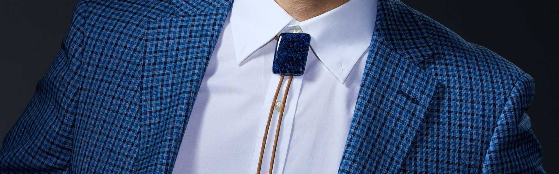 Modern blue dichroic glass bolo tie worn with white dress shirt and blue checkered blazer, featuring brown leather cord with silver tips